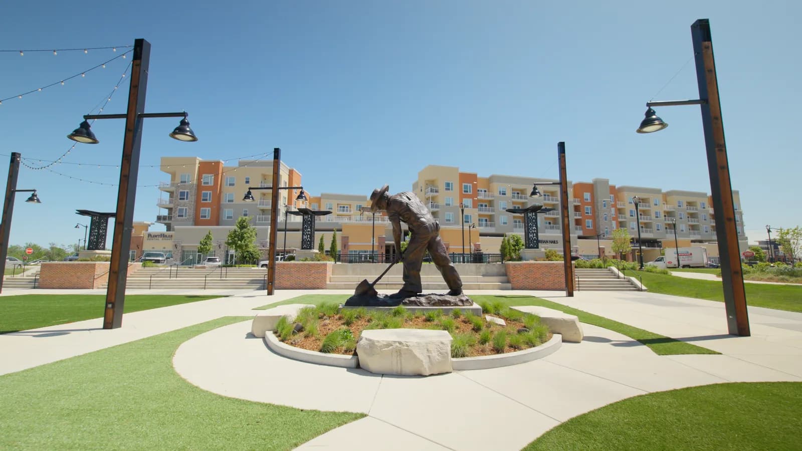 Bronze statue on a plaza with five-story apartment buildings and string lights behind it